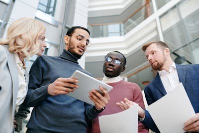 A diverse group of four professionals, two men and two women, are gathered closely, looking at a tablet held by one of the men. They appear to be in a modern office or atrium.