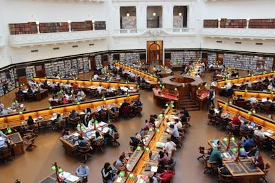 An overhead view of a grand, multi-tiered library reading room filled with numerous people studying at desks, surrounded by tall bookshelves.