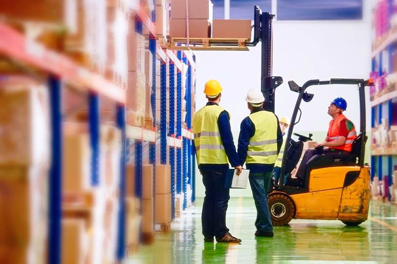 Three warehouse workers in safety vests and hard hats, with one operating a forklift to lift pallets of boxes, and two others observing.
